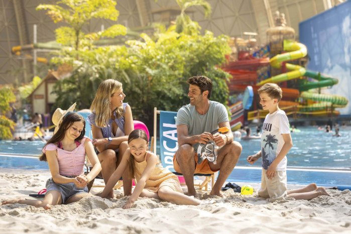 Familie mit drei Kindern auf Liegestühlen am Sandstrand vor Pool mit Rutschen und Palmen.