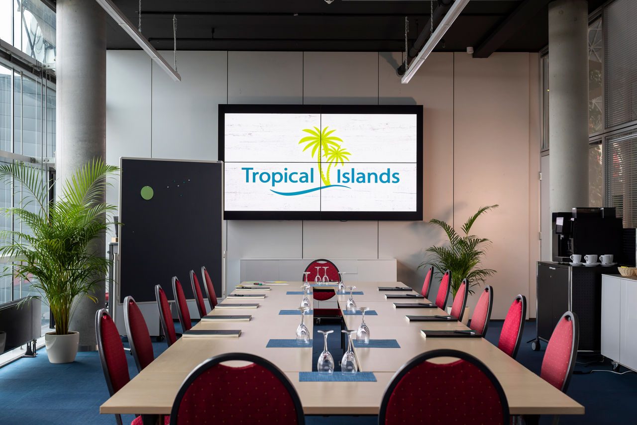 A bright conference room with rows of red chairs, a projection screen, large windows and plants.