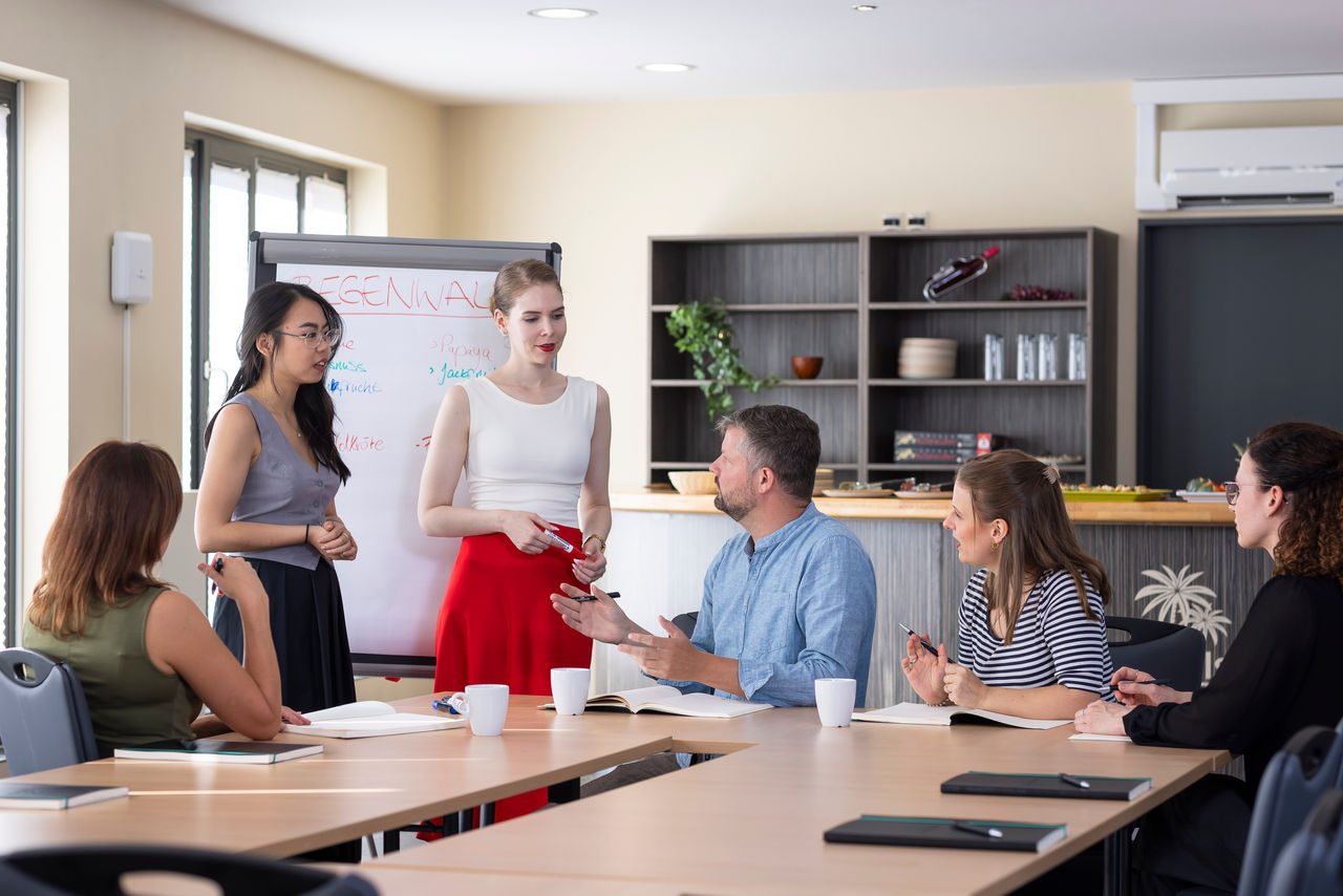 Six people/colleagues engaged in casual conversation during a conference, two standing in front of a flipchart