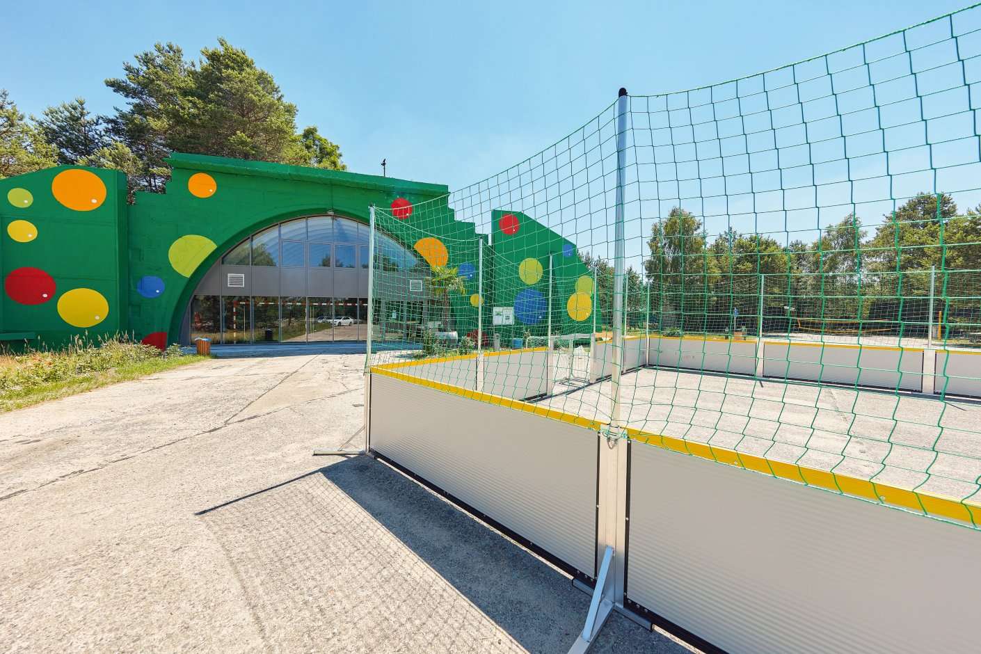 Outdoor-Sportanlage mit grünem Gebäude, bunten Dekoelementen und Netzbereich für Aktivitäten – umgeben von Natur.