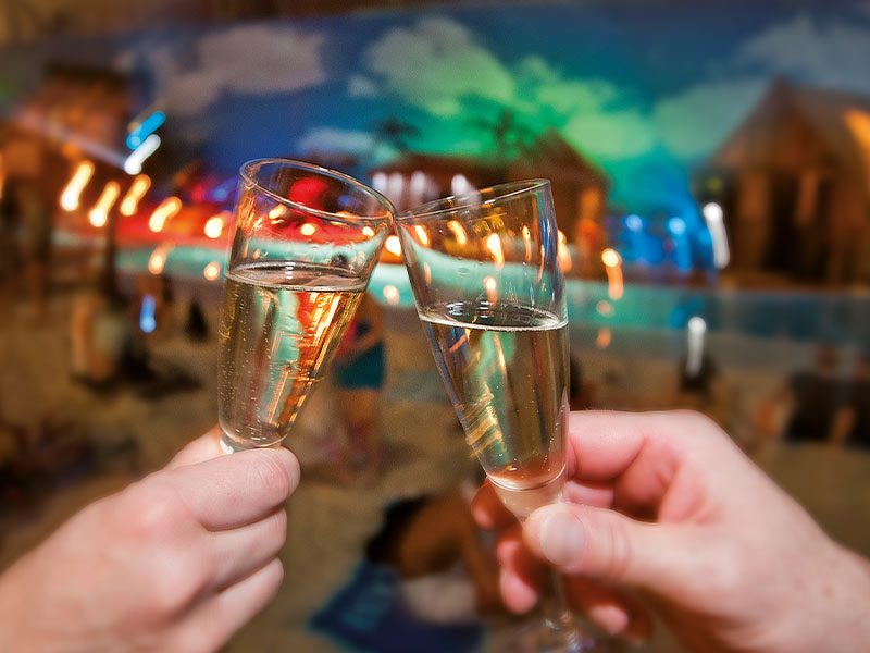 Zwei Sektgläser stoßen an, im Hintergrund etwas verschwommen die Südsee mit festlicher, bunter Beleuchtung.