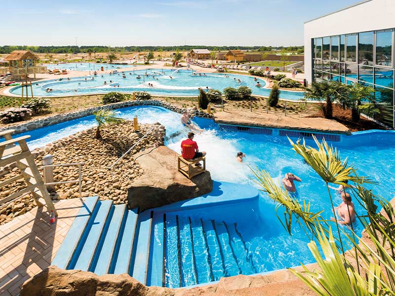 Outdoor area AMAZONIA with several pools, Whitewater River and bathers. A lifeguard sits on a chair. Surrounded by palm trees and plants.