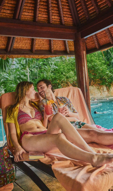 Mann und Frau mit Cocktails in den Händen liegen in einem Cabana an der Lagune und schauen sich lächelnd in die Augen.