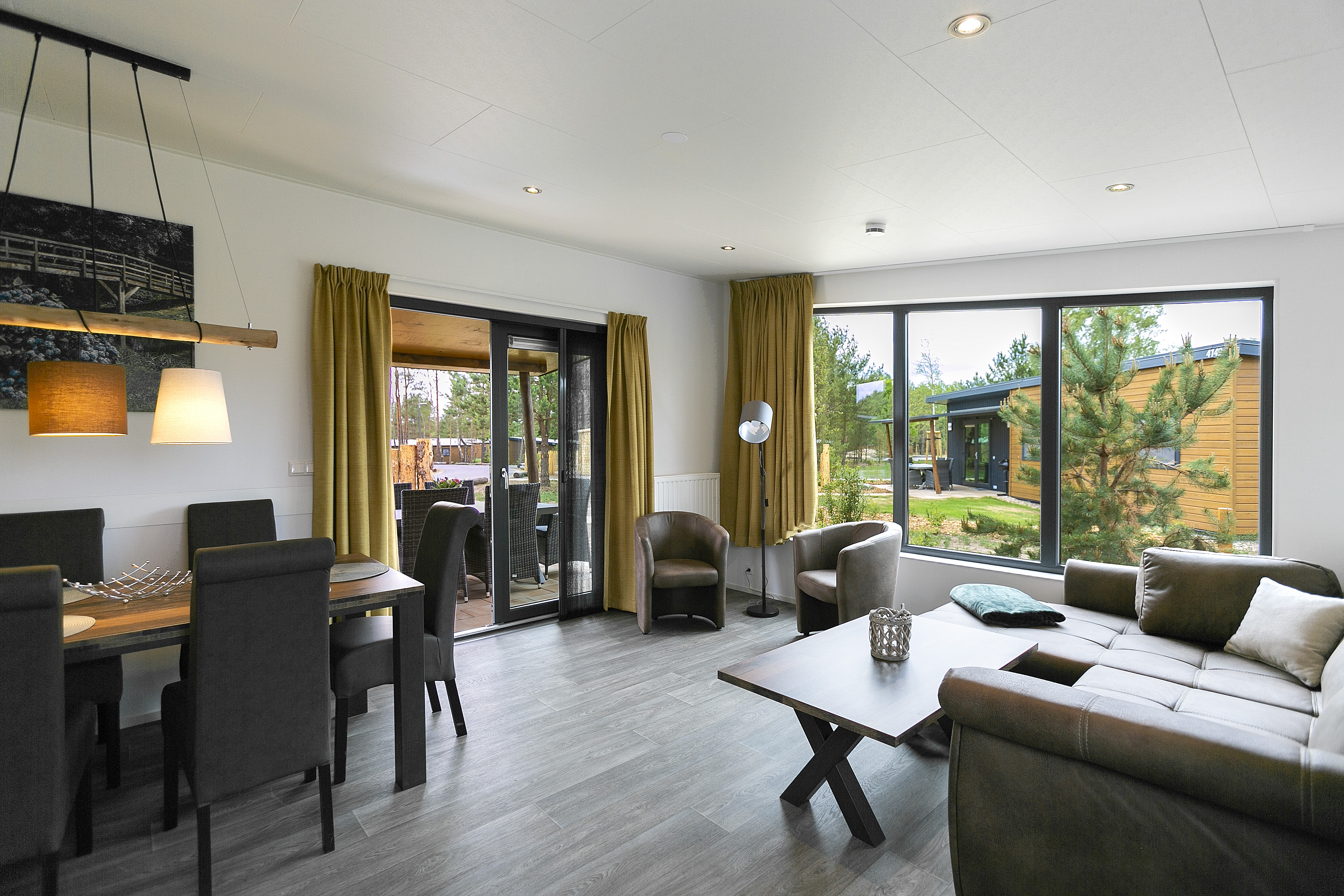 The interior of a Sunrise Home, furnished in a modern and stylish manner. On the left is a dining table with chairs, on the right is a seating area, and behind it are two large windows with mustard yellow curtains. 