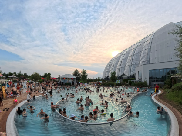 AMAZONIA Pool mit Menschen, einer Bühne im Hintergrund und dem Dome inklusive Sonnenschein und Wolken.
