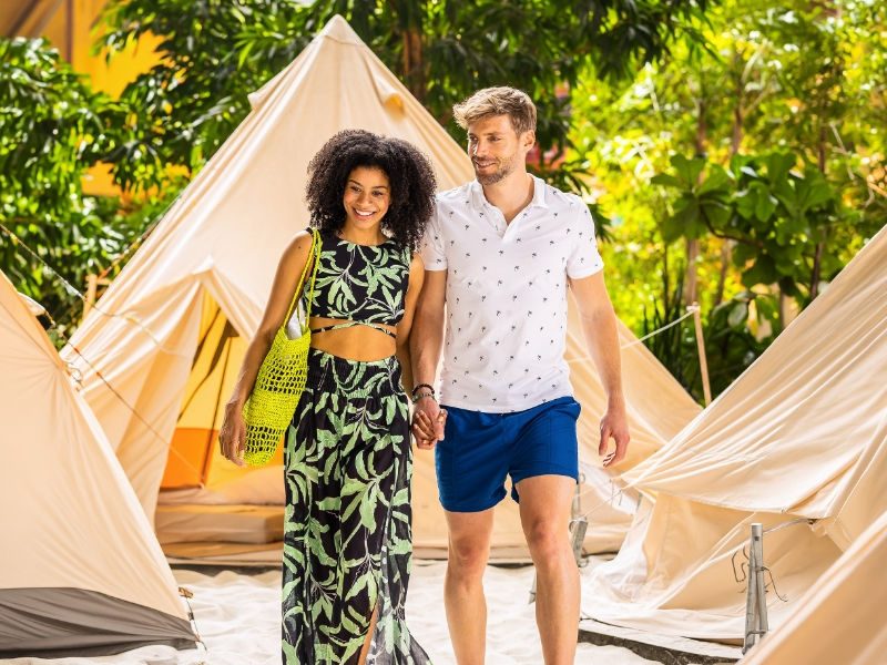Guests walking between light-colored tent accommodations at the Tropical Islands, surrounded by lush tropical greenery and warm sunlight.