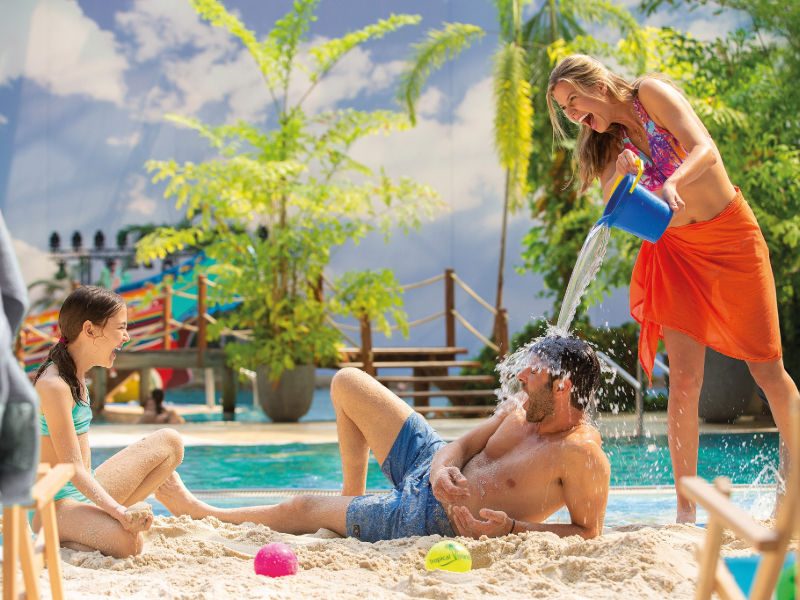 Familie sitzt im Sand, Frau kippt Mann Wasser über den Kopf.