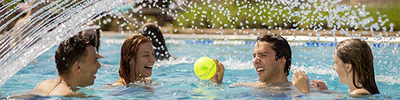 Gruppe junger Menschen spielt mit einem Ball im Pool, während ein Wasserstrahl über sie spritzt.
