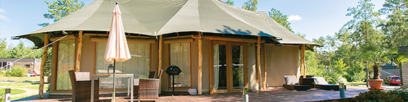 Großes Luxus-Zelt mit Holzterrasse, Gartenmöbeln und umgebender Begrünung unter leicht bewölktem Himmel.