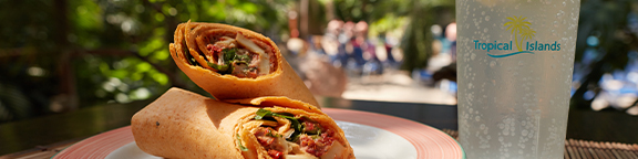 Zwei sonnenbeschienene Wraps schön angerichtet, daneben ein eisgekühltes Getränk in einem Becher mit Tropical Islands Aufschrift, im Hintergrund die Lagune.