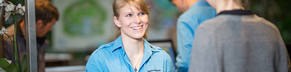 Person im blauen Shirt hinter einem Tresen im Gespräch mit einer Person im grauen Pullover; weitere Menschen und Pflanzen im Hintergrund.