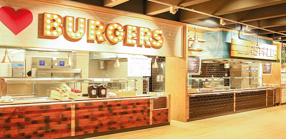 Ein heller Foodcourt mit zwei modernen Essensständen, darunter einer mit dem Schild „I ♥ BURGERS“.