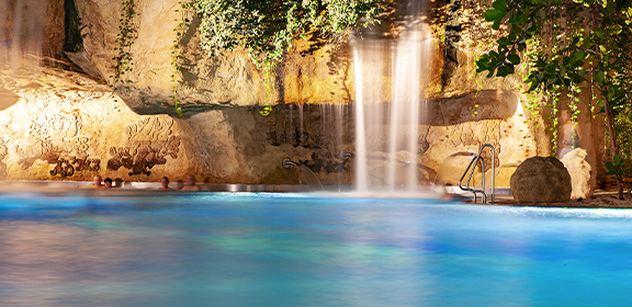 Ein Wasserfall fließt in eine klare, blaue Lagune. Umgeben von Felsen und grüner Vegetation. Rechts ist eine Badeleiter zu sehen