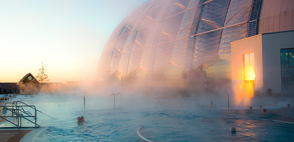 Ein Außenpool mit dampfendem Wasser, mehreren Personen und einer großen Glaskuppel im Hintergrund.