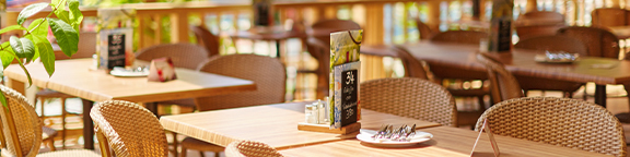 Restaurantbereich mit Holztischen, Korbstühlen und Speisekarten auf den Tischen.