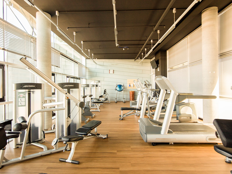 Fitness-Club mit Holzfußboden, Trainingsgeräten und großen Fenstern.