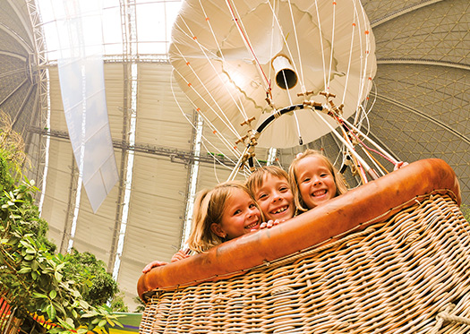 Ballonkorb mit drei Kindern unter einer großen Dachkonstruktion, umgeben von grünen Dekorationen.