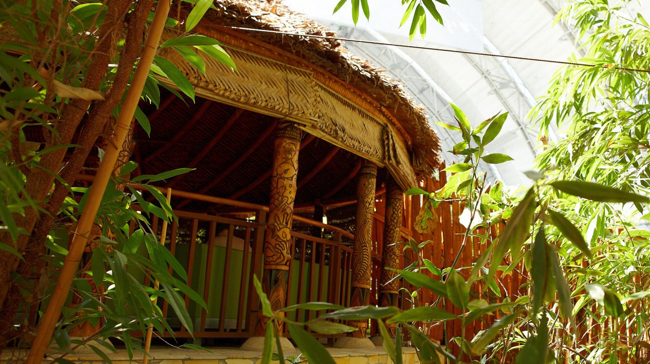Eine traditionelle Samoa-Fale Wohnhütte mit Strohdach und geschnitzten Holzpfeilern, umgeben von grünen Pflanzen.