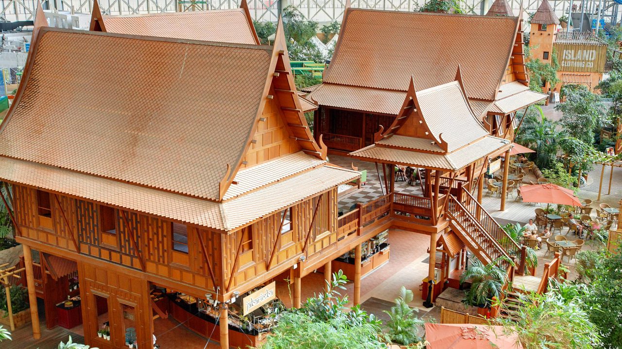 Traditionelle Thai-Häuser auf Stelzen mit verzierten Dächern im Tropendorf, umgeben von grünen Pflanzen.