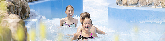 Drei Kinder treiben im Strömungskanal Whitewater River zwischen blauen Wänden und Steindekorationen.