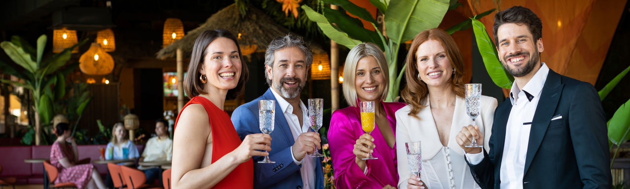 3 Frauen und 1 Mann in farbiger Business-Kleidung lächeln in die Kamera. In den Händen halten sie Trinkgläser.