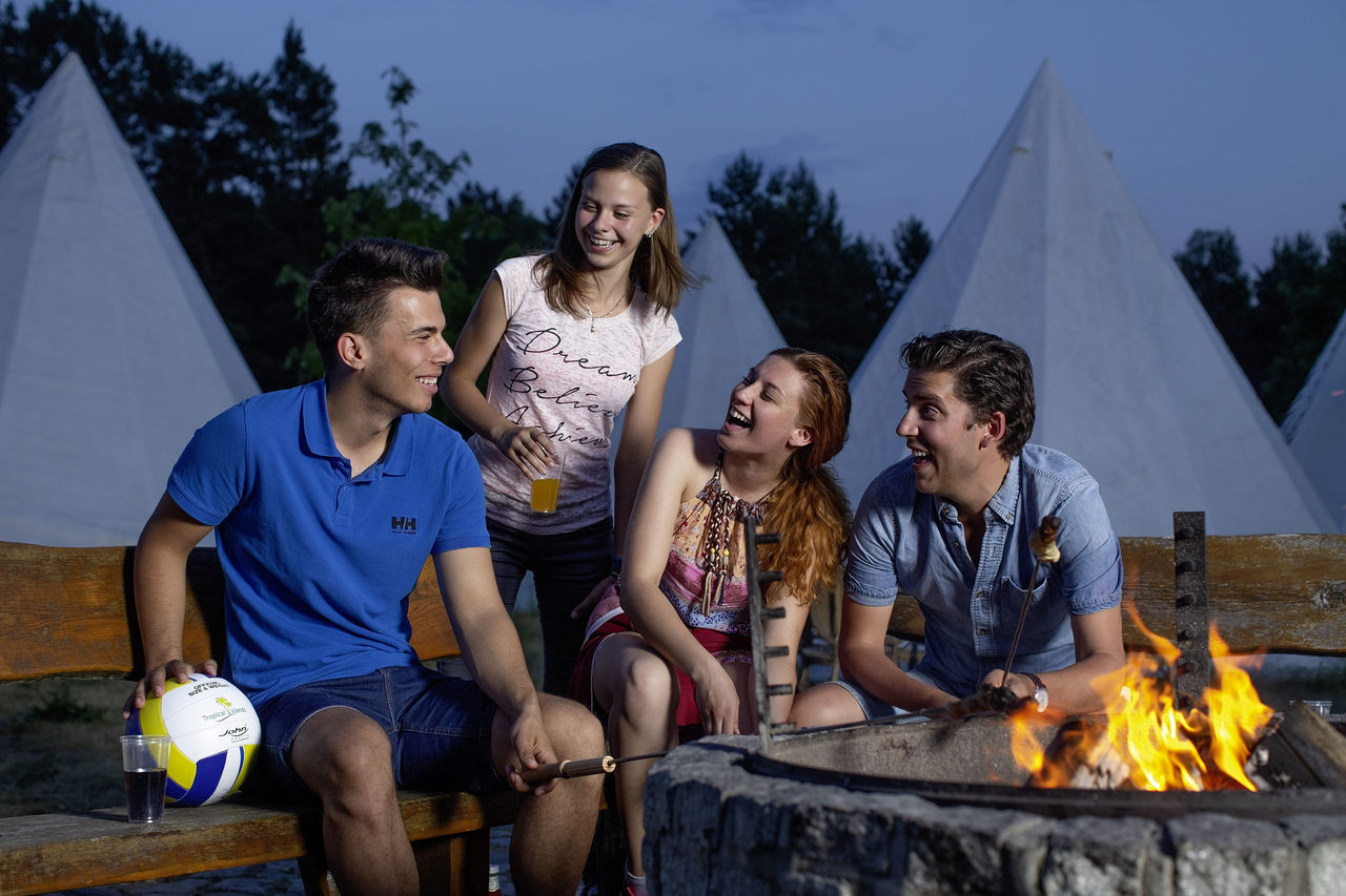 Gruppe lachend an einem Lagerfeuer, im Hintergrund weiße Tipi-Zelte.