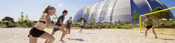 Sand-Volleyballfeld mit gelbem Netz und blauen Pfosten, umgeben von Bäumen und gelben Gebäuden.