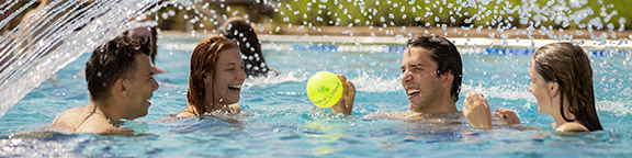 Gruppe junger Menschen spielt mit einem Ball im Pool, während ein Wasserstrahl über sie spritzt.
