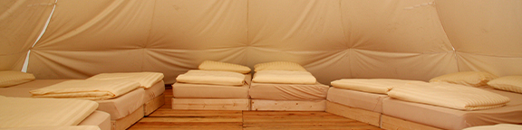 Innenansicht eines Zeltes mit mehreren Betten in einer Reihe, beige Zeltwände und einfache Ausstattung.