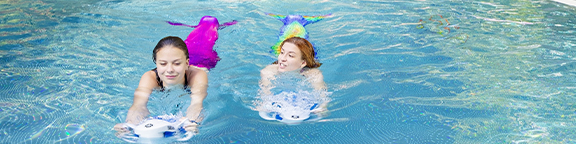 Zwei Personen mit Meerjungfrauenflossen fahren nebeneinander mit Unterwasser-Scootern im Pool.