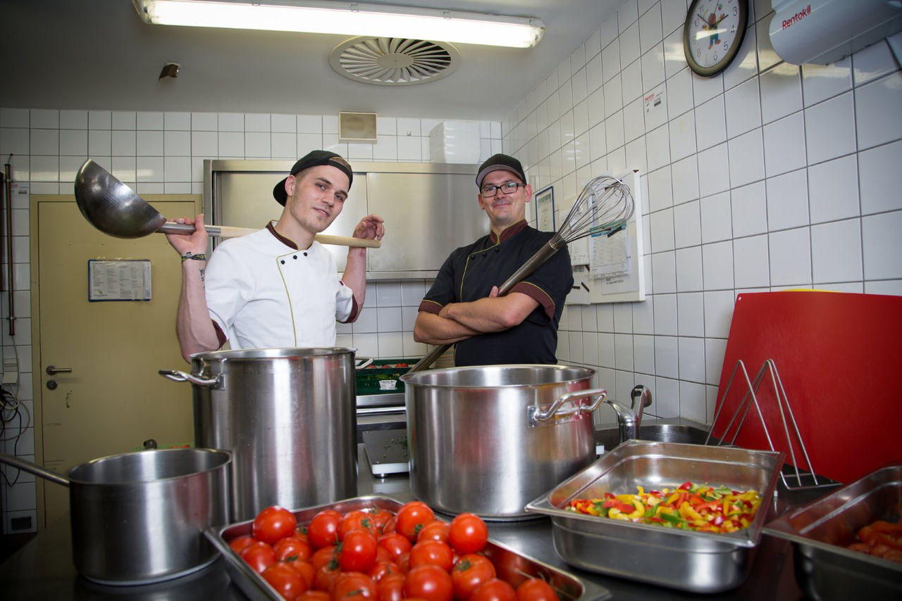 Zwei Personen stehen in der Küche mit Kochutensilien, großen Kochtöpfen und Gemüse.