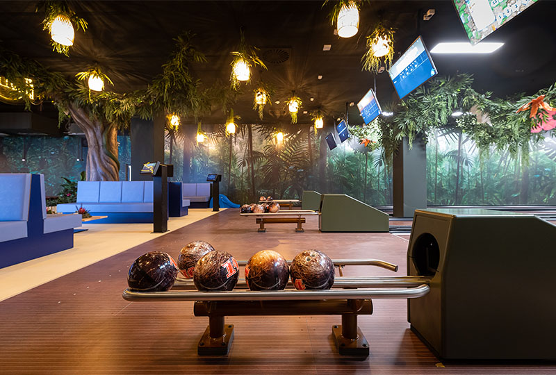 Eine tropisch gestaltete Bowlingbahn mit mehreren Bahnen, Dschungeldekoration und blauen Sitzbereichen.