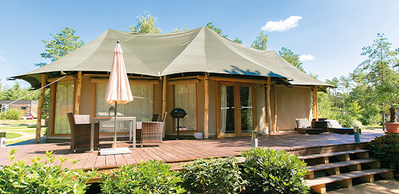 Großes Luxus-Zelt mit Holzterrasse, Gartenmöbeln und umgebender Begrünung unter leicht bewölktem Himmel.