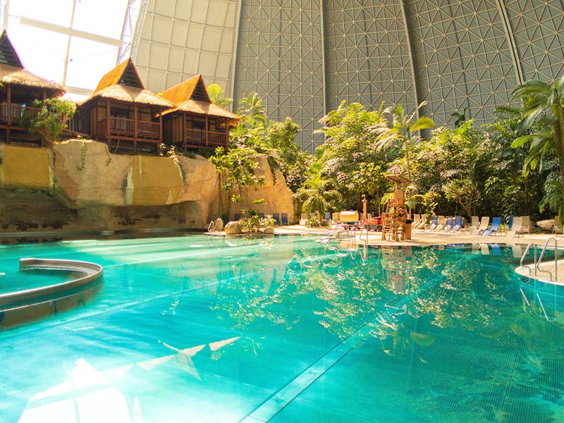 Die Lagune: Großer Pool mit Palmen, Glasdach und strohgedeckten Hütten in tropischer Umgebung.