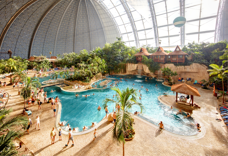 Tropische Badelandschaft unter einer großen Indoor-Kuppel mit türkisfarbenen Pools, Palmen, Wasserattraktionen, Liegebereichen und zahlreichen Gästen in sommerlicher Urlaubsatmosphäre.