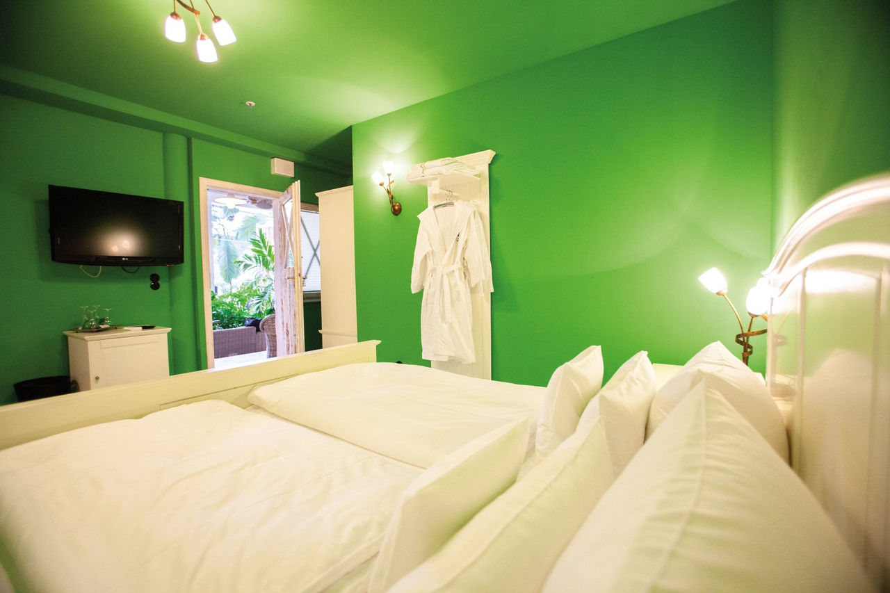 Helles Hotelzimmer mit grünen Wänden, großem Bett, TV, offenem Türzugang ins Grüne und Bademantel an der Wand.