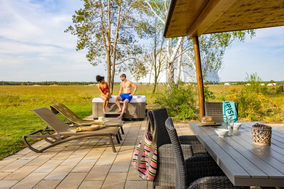 Terrasse mit Sitzgruppe, Liegen und Whirlpool vor Wiese und Bäumen bei klarem Himmel.