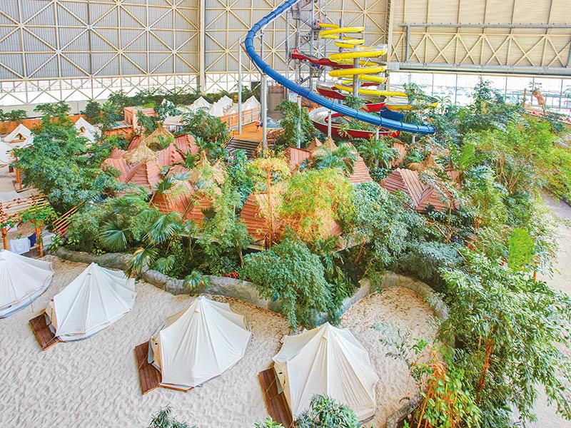 Indoor-Resort mit weißen Zelten auf Sand, tropischer Bepflanzung und Wasserrutschen-Turm.
