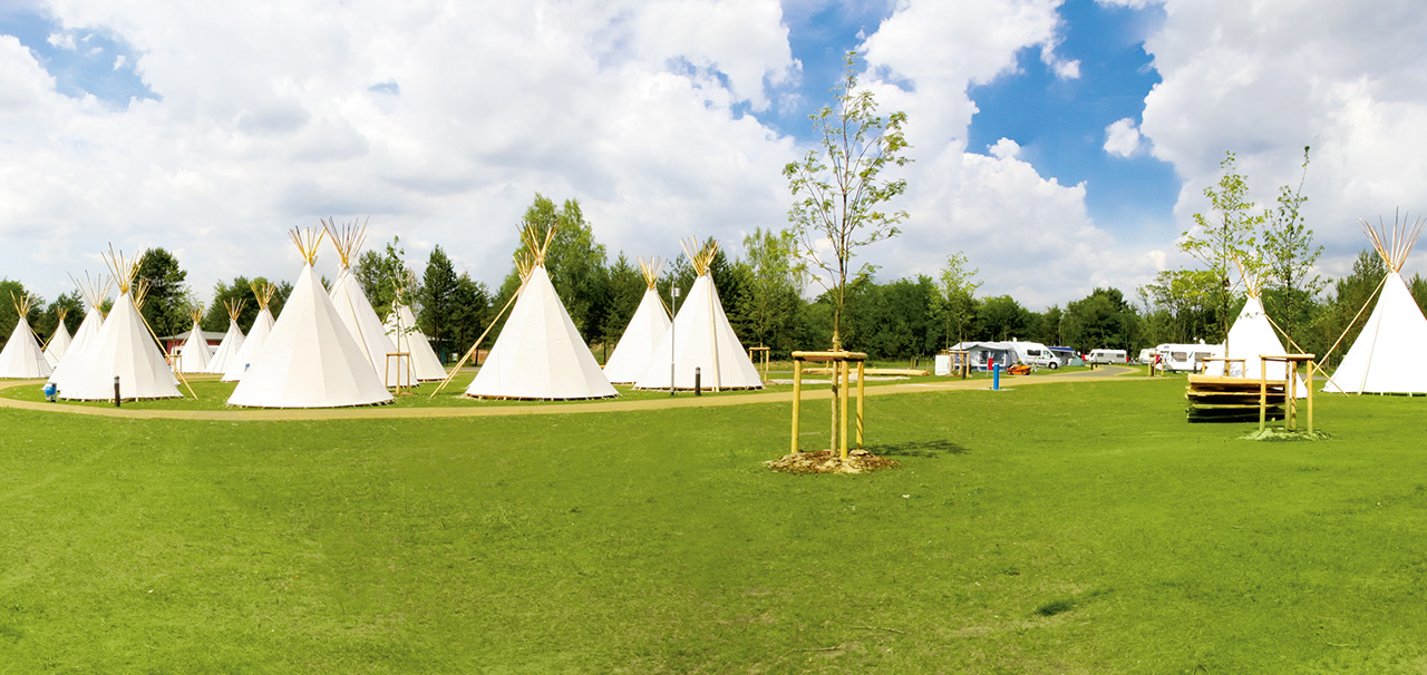 Weiße Tipis auf einer Wiese unter teils bewölktem Himmel, umgeben von Bäumen.