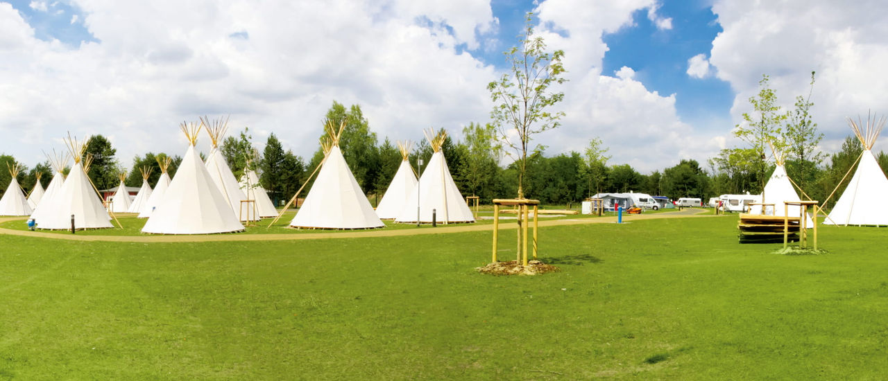 Weiße Tipis auf einer Wiese unter teils bewölktem Himmel, umgeben von Bäumen.