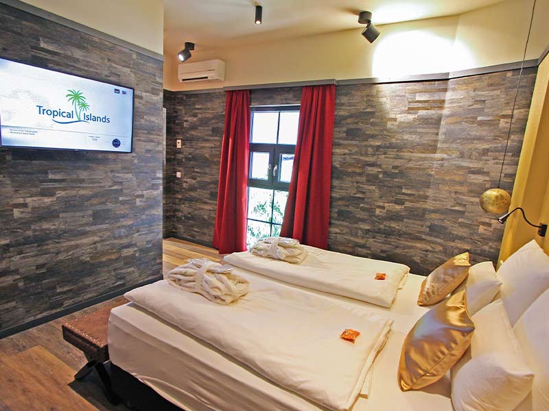 Sky Suite mit zwei Doppelbett, Steinwand, rotem Vorhang und TV mit Tropical-Islands-Logo.