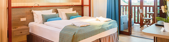 Bright hotel room with large bed, blue accents and access to the balcony with wooden furniture and a view of the greenery