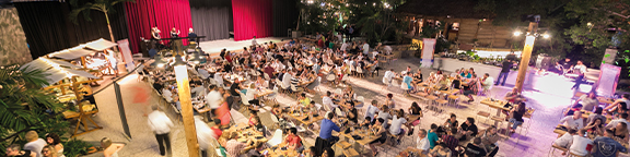 Large event with lots of people at tables, stage with red curtains in the background, festive lighting.