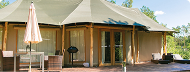Large luxury tent with wooden terrace, garden furniture and surrounding greenery under a slightly cloudy sky.