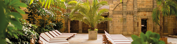 Sun-drenched sun loungers in the sauna area, surrounded by palm trees and greenery, with the walls of a temple in the background.