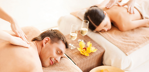 Two people are getting back massages on tables, with champagne glasses and a flower beside them.