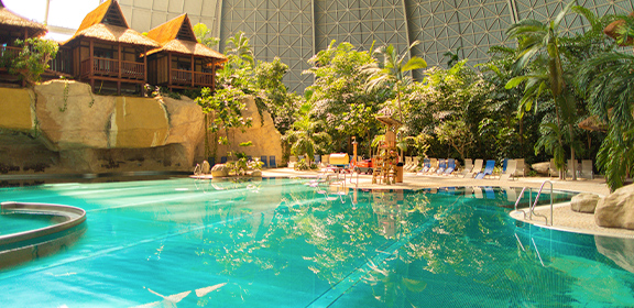 Lagoon with swimming pool, surrounded by tropical plants and sun loungers and wooden huts.