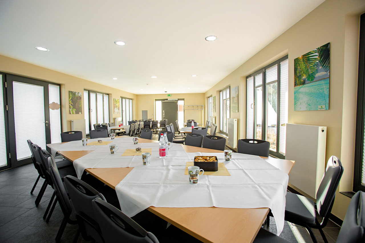 A bright room with laid tables, white tablecloths, pastries and glass jars, surrounded by black chairs.