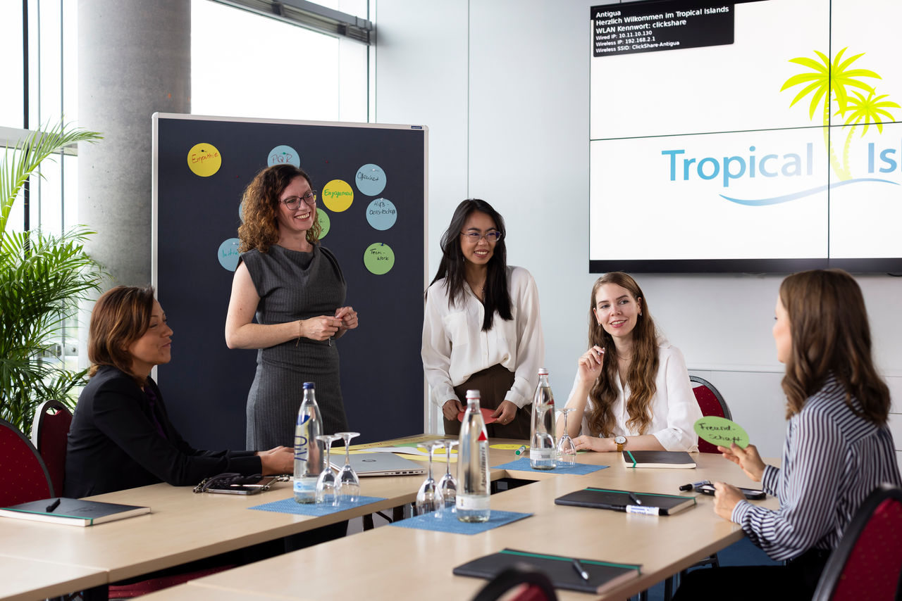 Five women/colleagues talking during a conference, with a screen and a pinboard with colorful notes in the background.
