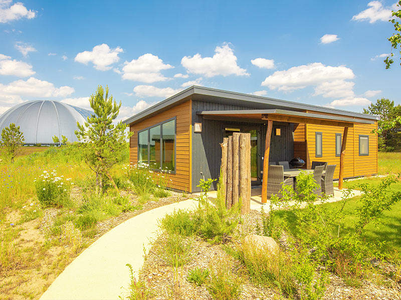A Sunrise Home with wooden exterior and large windows stands on a grassy area, with a path to the door and a dome in the background.